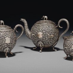 Comprising a tea pot, a creamer and a sugar bowl with cover, the teapot and sugar bowl with repoussé decoration depicting felines chasing and pouncing on their prey amongst scrolling flowers and foliage, the creamer with a cheetah, a parakeet and an elephant on similar ground, each with central shield cartouche, a handle modelled on a snake and with finials in the form of an elephant, each on four feet and marked V.K.